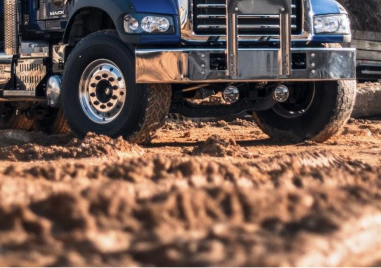 Close-up of a blue Mack truck's front grille and wheels parked on muddy terrain.