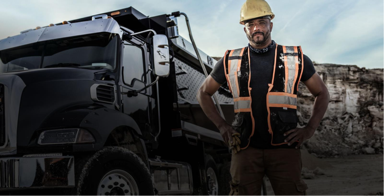 Construction worker in a safety vest and hard hat standing confidently next to a black Mack dump truck at a job site