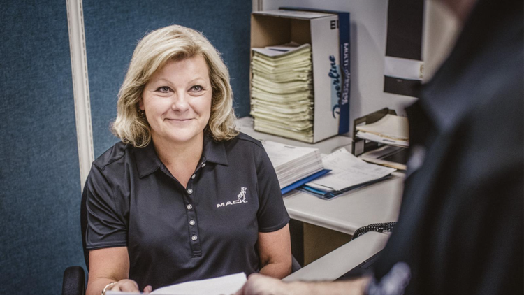A Mack Trucks employee in a branded polo shirt assisting a customer, with warranty documents on the desk and organized files in the background