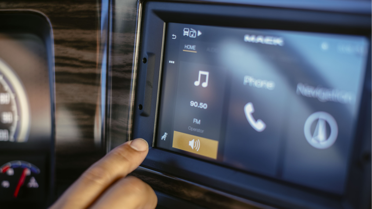 Close-up of a Mack truck's infotainment touchscreen displaying music, phone, and navigation options, with a hand interacting.