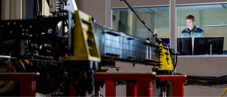 A man working at a computer in a control room, overlooking heavy-duty equipment being tested on a platform inside a workshop