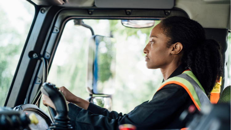 Driver in a safety vest seated in a Mack truck cab, looking ahead through a large side window.