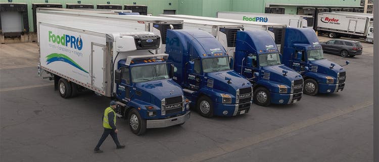 Four blue Mack trucks with 'FoodPRO' trailers parked at a distribution center.