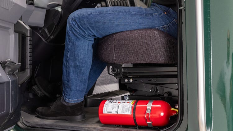 A driver seated in the cab of a Mack MD Series truck with a fire extinguisher secured under the seat.